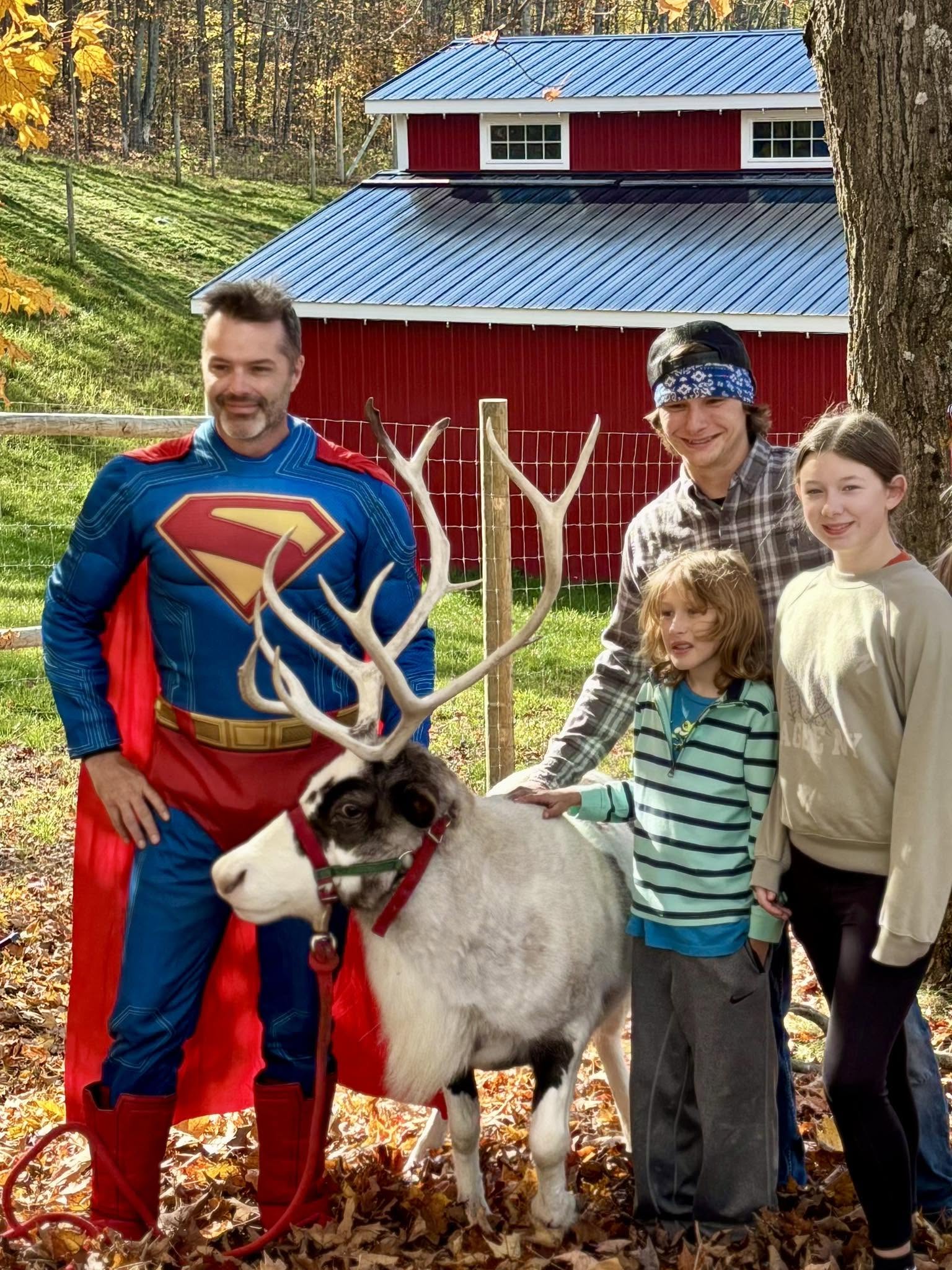 Superman posing with a reindeer and kids by the red barn at Halloween On The Farm, Wilderness Valley Farm near Gaylord, Michigan